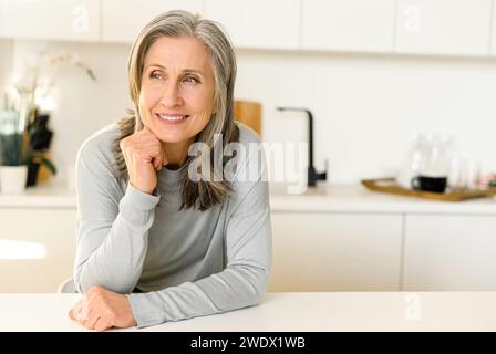Una donna anziana tranquilla con i capelli grigi trascorre il tempo libero a casa, ispirata a una donna dai capelli grigi che sogna ad occhi aperti, guarda lontano perduta nei pensieri, seduto alla scrivania in cucina Foto Stock