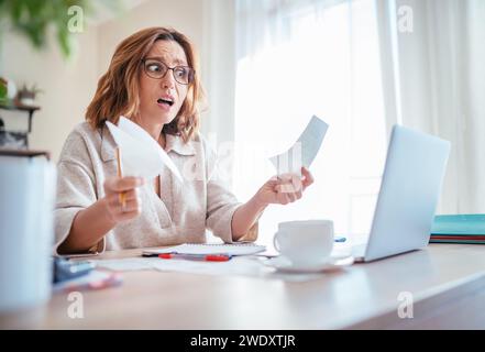 Bella donna di mezza età con gli occhiali ha sorpreso molto le spese familiari più alte osservando le bollette di carta e l'ufficio domestico del portatile. Problemi delle piccole imprese Foto Stock