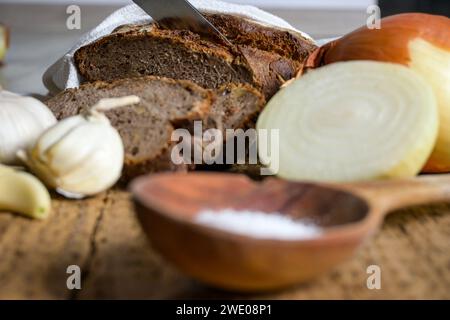 pane su un panno con un coltello, aglio, cipolla, sale e un cucchiaio di legno su uno sfondo di legno. Foto Stock