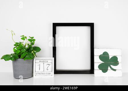 Montatura nera con motivo St Patricks Day su uno scaffale bianco. Pianta di Shamrock, calendario e segno in legno shabby chic. Cornice verticale contro una wa bianca Foto Stock