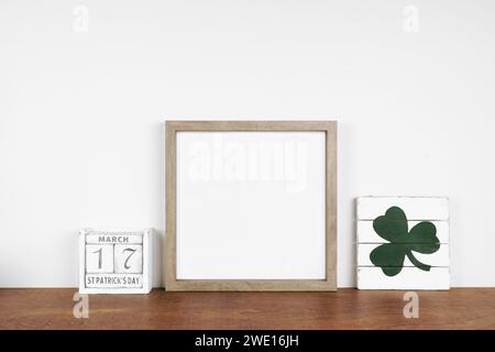 Montatura in legno con decorazioni St Patricks Day su un ripiano in legno. Cartellone e calendario in legno squallido ed elegante. Cornice quadrata contro una parete bianca. Copia spazio. Foto Stock