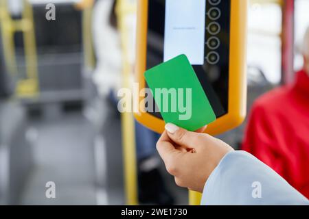 Coltura di mano femminile che utilizza il validatore per il pagamento durante l'imbarco nei trasporti pubblici. Primo piano della mano nella custodia blu con la carta verde che paga il viaggio con il trasporto. Concetto di dispositivo elettronico. Foto Stock