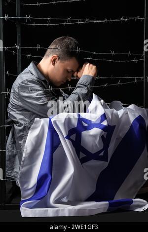 Giovane ebreo con bandiera di Israele che prega dietro filo spinato su sfondo nero. Giornata internazionale della memoria dell'Olocausto Foto Stock