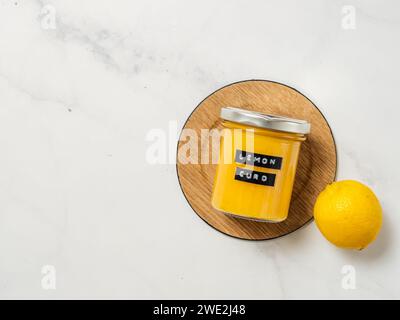 Deliziosa cagliata al limone fatta in casa. Vaso in vetro con cagliata di limone su fondo in marmo bianco. Etichetta adesiva nera con testo Lemon Curd sul vaso. Vista dall'alto o disposizione piatta. Copia spazio Foto Stock