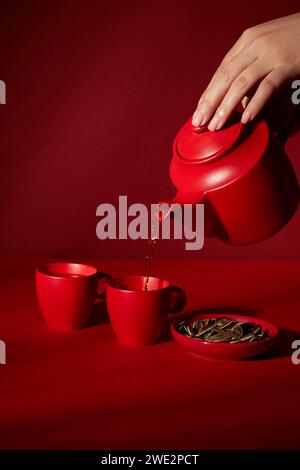 Mano femminile che tiene una pentola rossa e versa il tè in tazza. Set di pentola rossa e tazza decorata su sfondo rosso scuro. Sfondo astratto minimo per gli annunci pubblicitari Foto Stock