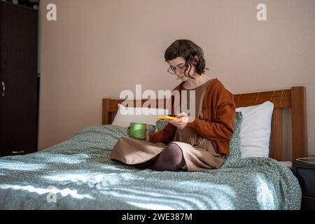 Ragazza adolescente che legge messaggi scorrendo i social media nello smartphone bevendo tè a letto. Dipendenza telefonica Foto Stock