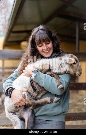 Felice giovane contadina che abbraccia una piccola pecora in una fattoria rurale biologica. Benessere e cura degli animali in un fienile. Foto Stock