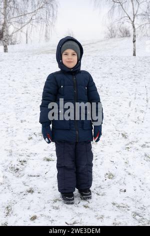 Un bambino di sei anni in tuta blu d'inverno in un parco innevato Foto Stock