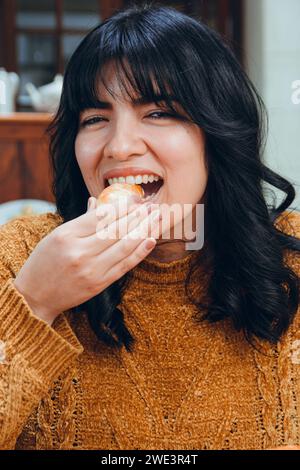 Immagine verticale di una giovane latina con capelli neri e vestiti marroni, happy sitting in un ristorante che mangia tequenos guardando la macchina fotografica, tradizionale Venezuela Foto Stock