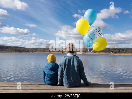 una donna e un bambino siedono con la schiena vicino a un lago in una giornata di sole, tenendo in mano palloncini gialli e blu Foto Stock