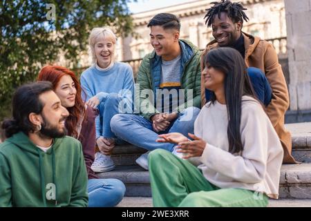 Gruppo di giovani adulti felici di varie etnie impegnati in una vivace discussione sui gradini del campus, condividendo un momento di gioia. Foto Stock