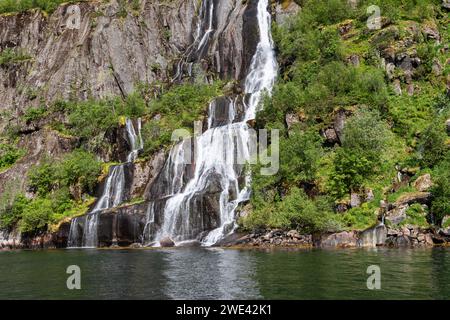 L'acqua illuminata dal sole sgorga lungo una ripida parete rocciosa nelle tranquille acque di Trollfjorden a Lofoten, Norvegia, incorniciate da una vegetazione lussureggiante Foto Stock