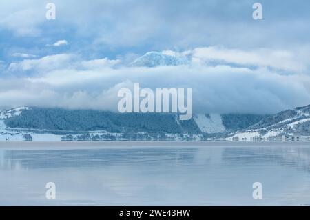 Un tranquillo paesaggio invernale mostra una montagna innevata ricoperta di nuvole, che si riflette perfettamente nelle calme acque del mare. Foto Stock