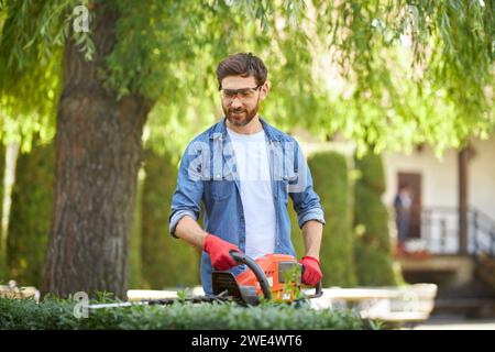 Giardinaggio maschio con barba sorridente che taglia il cespuglio in legno di bosso con tagliasiepi nelle giornate di sole. Vista frontale del tuttofare che utilizza attrezzature per il giardinaggio per modellare la siepe. Concetto di lavoro stagionale, hobby. Foto Stock