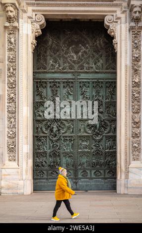 Una donna vestita con un cappotto giallo, un cappello e delle scarpe passa davanti a porta maggiore, la porta principale del Duomo di Milano, Italia. Foto Stock