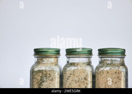 Tre vasetti di aglio, sale ed erbe aromatiche condite con spezie sono vicini di fila. Contenitore in vetro con coperchio chiuso in alluminio verde su sfondo grigio, ricicla Foto Stock