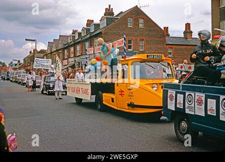 Sfilata annuale del Carnevale Whitsun di Watford che procede lungo St Albans Road, Watford, Hertfordshire, Inghilterra, Regno Unito, 1967. uno Scammell giallo 'Townsman' è dotato di un autocarro a pianale con caricatore basso. Le donne con palloncini promuovono il servizio ferroviario British Railways Londra-Manchester. La Scammell Trucks era un'azienda locale di Watford. Davanti al Tuck c'è un altro veicolo in processione che promuove il servizio di emergenza volontario. Dietro il camion, un uomo tiene un cartello davanti a un minivan con la scritta: PDSA, è necessario il tuo aiuto: Una fotografia d'epoca degli anni '1960. Foto Stock