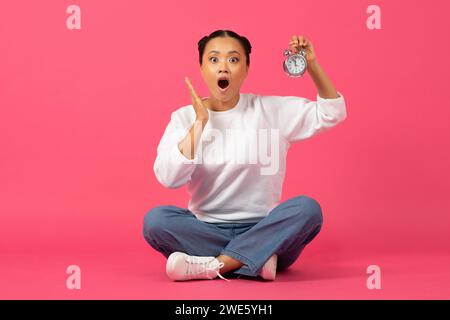 Una giovane donna asiatica scioccata seduta sul pavimento e con la sveglia in mano Foto Stock