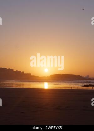 Le onde si infrangono su una roccia al tramonto su una spiaggia sabbiosa con medina a sinistra, a Essaouira, Marocco, 23 gennaio 2024 Foto Stock