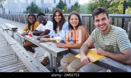 Gruppo di studenti multietnici seduti insieme fuori dall'università per svolgere un lavoro scolastico guardando la telecamera Foto Stock