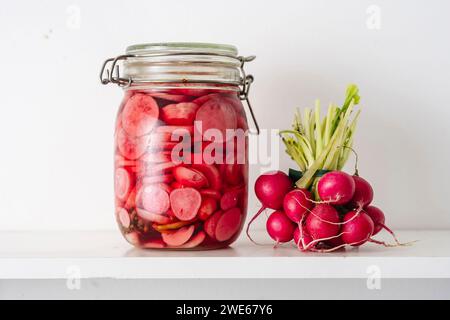 Barattolo con ravanelli sottaceto vicino alle verdure sullo scaffale a casa Foto Stock