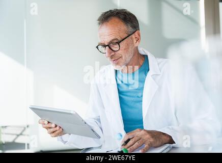 Medico sorridente che utilizza un tablet PC e seduto con il blocco note alla scrivania Foto Stock