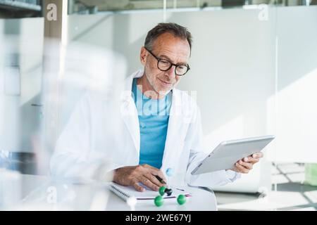 Un medico anziano sorridente che utilizza un tablet PC e si siede con un blocco note alla scrivania Foto Stock