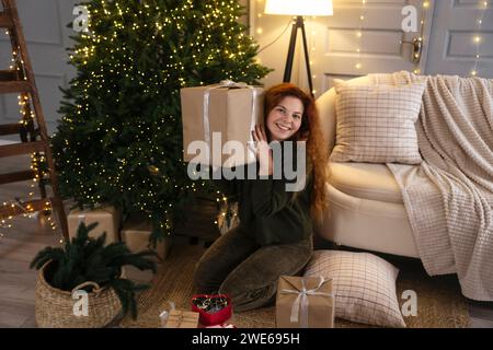 Donna felice che tiene una confezione regalo vicino all'albero di Natale a casa Foto Stock