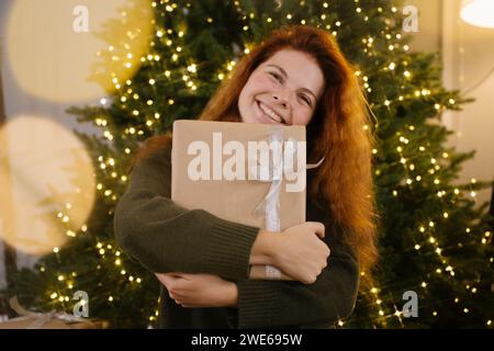 Donna sorridente con la testa rossa che tiene una confezione regalo vicino all'albero di Natale Foto Stock