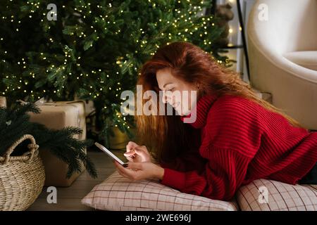 Felice donna rossa che usa il cellulare vicino all'albero di Natale a casa Foto Stock