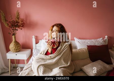 Donna con la testa rossa contemplativa che tiene una tazza di caffè a letto a casa Foto Stock