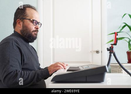 Uomo che suona pianoforte elettrico e vlogging con smartphone a casa Foto Stock