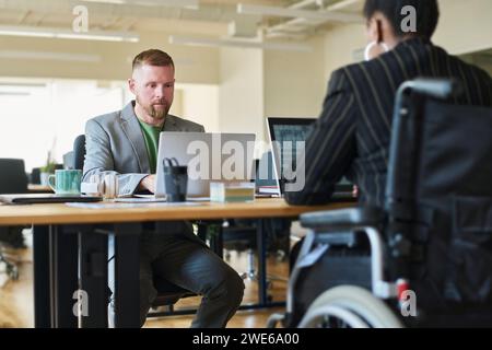 Donna d'affari in sedia a rotelle che lavora su un computer portatile con un collega alla scrivania in ufficio Foto Stock