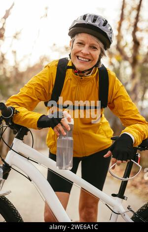 Donna felice che tiene una bottiglia d'acqua e si appoggia sulla mountain bike nella foresta Foto Stock