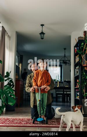 Padre sorridente che assiste il figlio a guidare lo scooter a casa Foto Stock