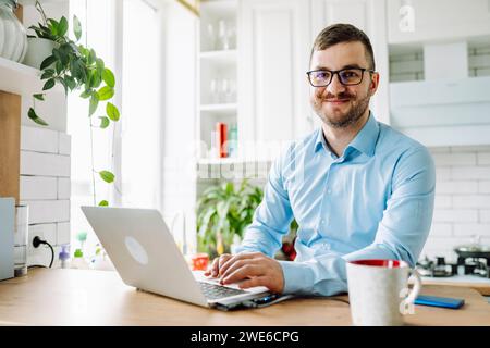 Freelance sorridente seduto con un computer portatile al tavolo in cucina Foto Stock