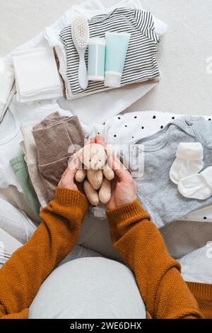 Donna incinta che tiene un orsacchiotto imbottito vicino ai vestiti a casa Foto Stock