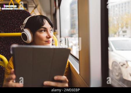 Donna che indossa cuffie wireless e si siede con un tablet PC in autobus Foto Stock