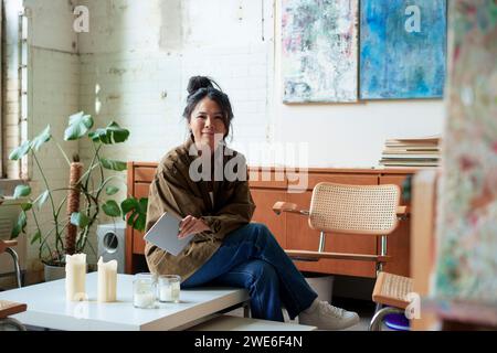 Artista sorridente con tablet PC seduto sul tavolino in officina Foto Stock