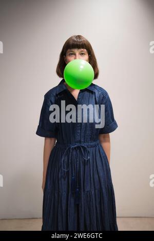 Giovane donna che soffia palloncino verde davanti al muro Foto Stock