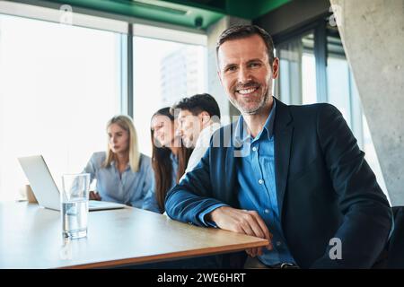 Uomo d'affari maturo sorridente seduto con colleghi che lavorano in background Foto Stock
