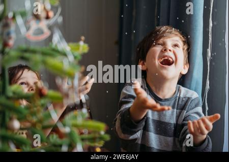 Ragazzi giocosi che prendono la neve finta vicino all'albero di Natale a casa Foto Stock