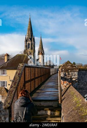 Persona che cammina su un vecchio ponte di legno verso una cattedrale con guglie sotto un cielo blu. Foto Stock