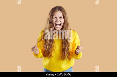 Frustrata emotiva arrabbiata triste urlando una giovane donna con pugni aggrappati su sfondo beige. Foto Stock