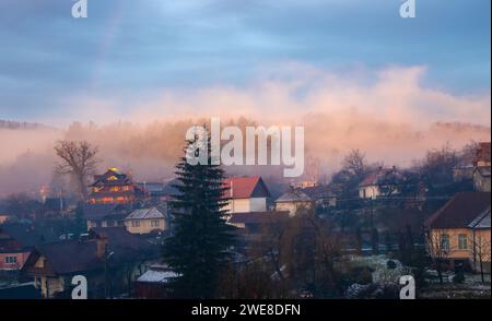 Un piccolo villaggio nebbioso avvolto dalla nebbia e circondato da alberi lussureggianti al mattino Foto Stock