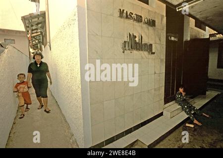 Una donna cammina con un bambino attraverso un vicolo sul lato di una moschea, dove un altro bambino è seduto alla sua porta d'ingresso; fotografato in un quartiere fitto vicino alla zona di Gegerkalong a Bandung, Giava Occidentale, Indonesia. Foto Stock