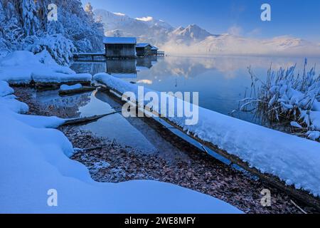 Boathouses, riflesso delle montagne nel lago, inverno, neve, sole, Lago Kochelsee, alta Baviera, Baviera, Germania, Europa Foto Stock