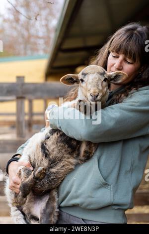 Felice giovane contadina che abbraccia una piccola pecora in una fattoria rurale biologica. Benessere e cura degli animali in un fienile. Foto Stock