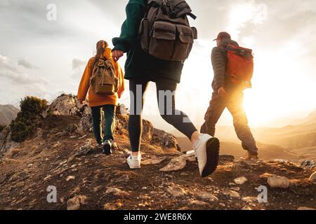 Tre giovani escursionisti con piccoli zaini camminano sulle montagne al tramonto. Foto ravvicinata delle gambe turistiche alla luce del tramonto Foto Stock