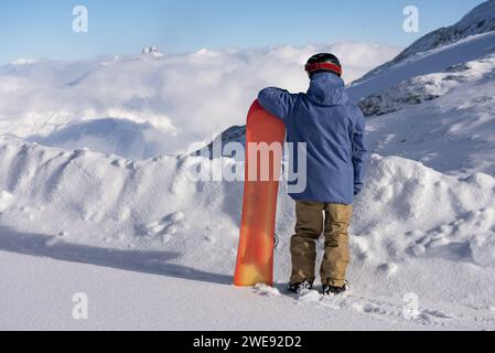 L'uomo tiene uno snowboard sullo sfondo delle alpi. Inverno nelle alpi francesi. Lo snowboarder si trova sulle alpi con uno snowboard. Foto Stock